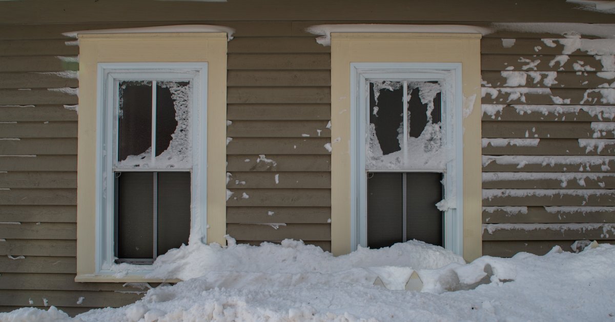 An exterior wall of a home clad with vinyl siding and featuring two windows. A large drift of white snow reaches the windowsills.