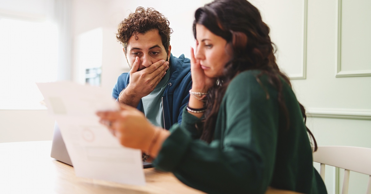 A young stressed couple looking at a bill while sitting at their kitchen counter. The man covers his mouth in shock.