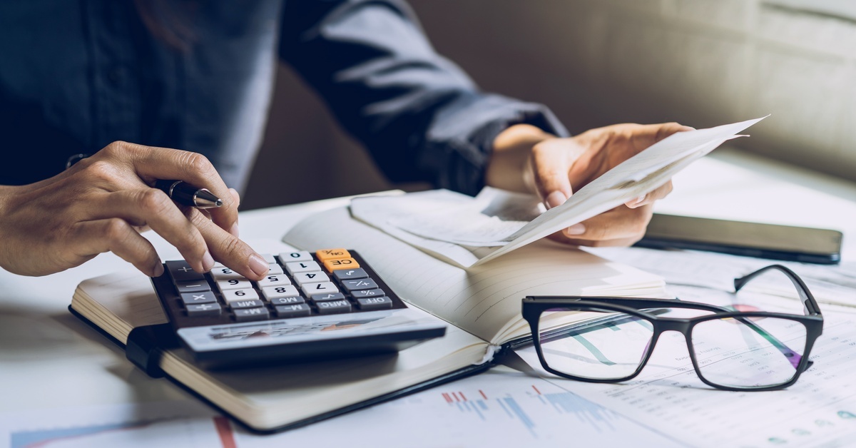 A person holding a bill in one hand and typing on a calculator with the other on a desk strewn with other documents. A person holding a bill in one hand and typing on a calculator with the other on a desk strewn with other documents.