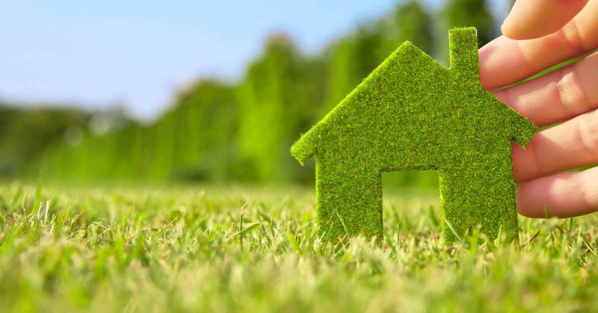 A hand holding up a small mossy cut-out of a home in a field of grass under a clear blue sky. A hand holding up a small mossy cut-out of a home in a field of grass under a clear blue sky.