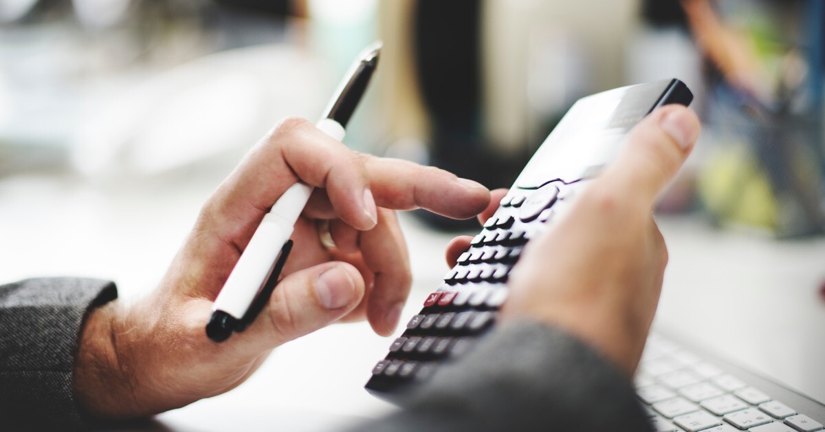 A close-up of a man holding a calculator in one hand while pressing its buttons and holding a pen with the other.
