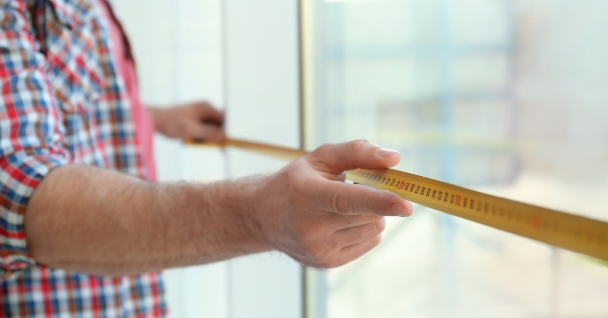 A man extending a yellow tape measurer to find the width of a window he's standing in front of.
