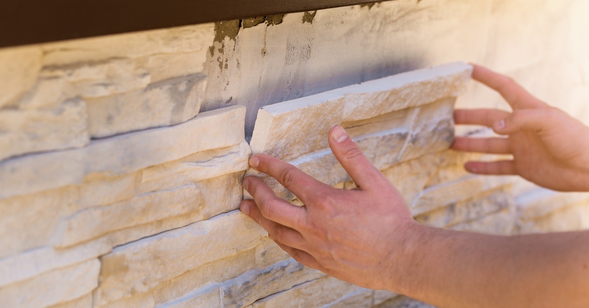A close-up of a person's hands placing a rectangle of white stone veneer onto a mortared decorative wall in progress. A close-up of a person's hands placing a rectangle of white stone veneer onto a mortared decorative wall in progress.