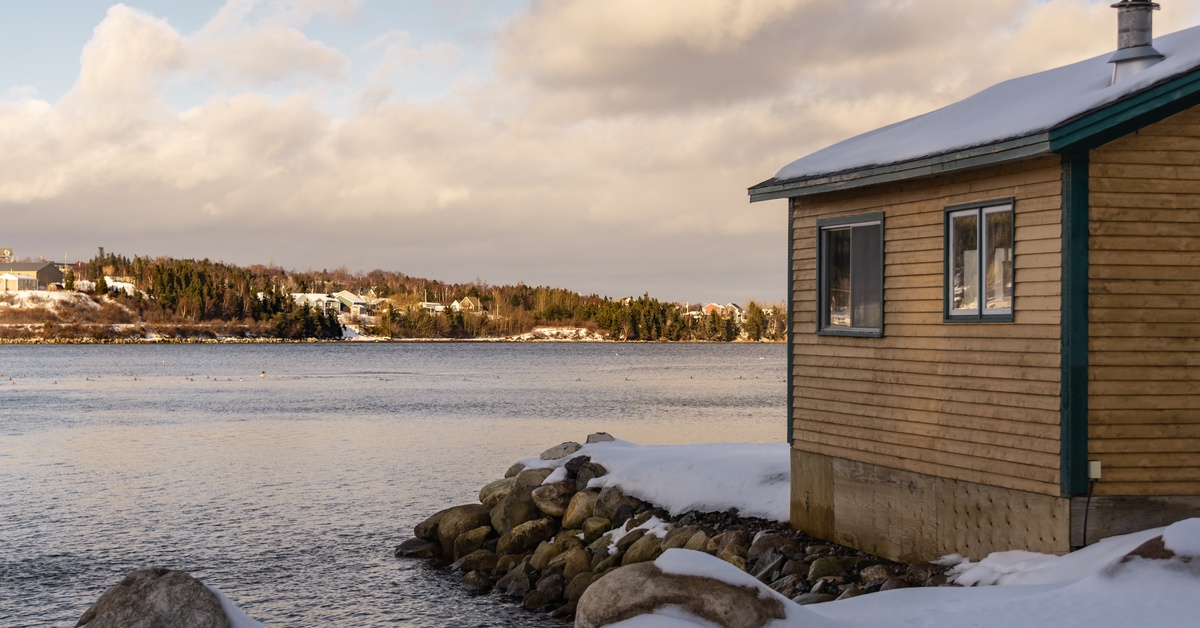 Beautiful Cabin Siding Ideas To Elevate Your Exterior A small cabin with plank siding and dark green trim facing a lake at dusk. The roof and ground are covered in snow.