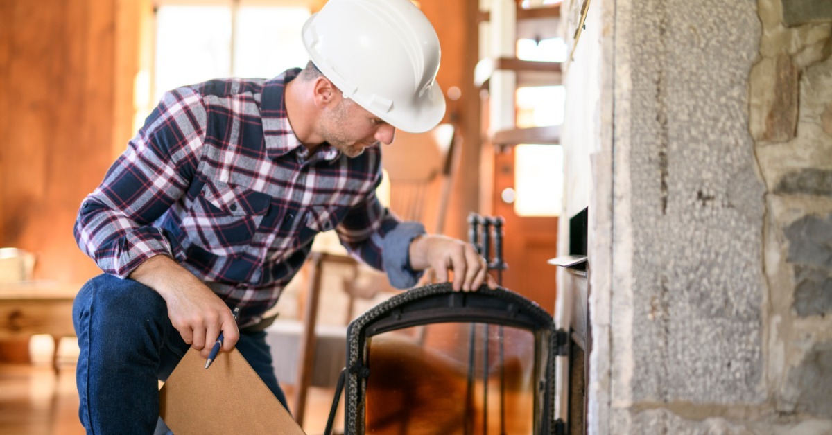 A contractor in a white hard hat checks a cabin's open glass fireplace door on a stone hearth while holding a clipboard.