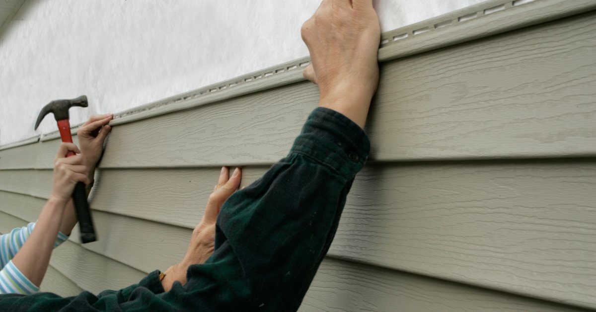 Two people are holding a piece of wood-textured vinyl siding against a house. One of them is hammering it in.